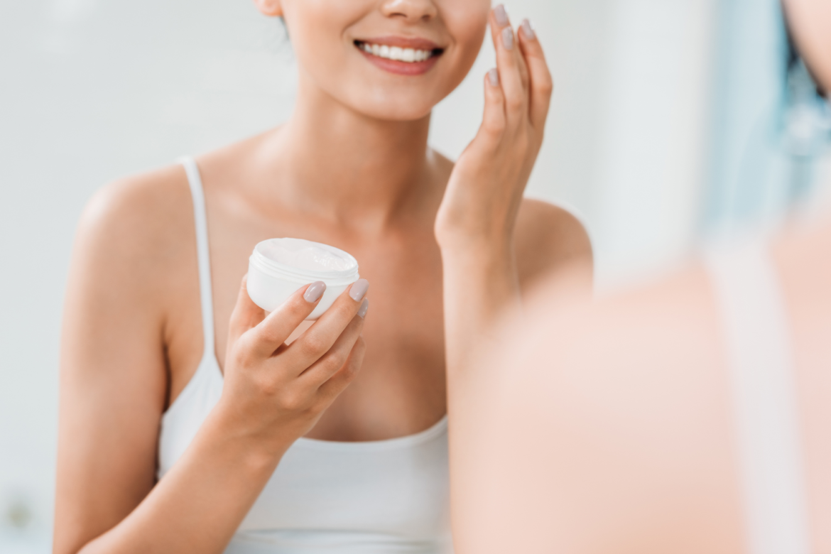 cropped shot of smiling girl holding container and applying face cream at mirror in bathroom