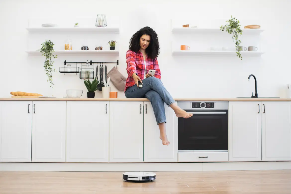 Young Woman in Kitchen Using Smartphone and Monitoring Robotic Vacuum Cleaner